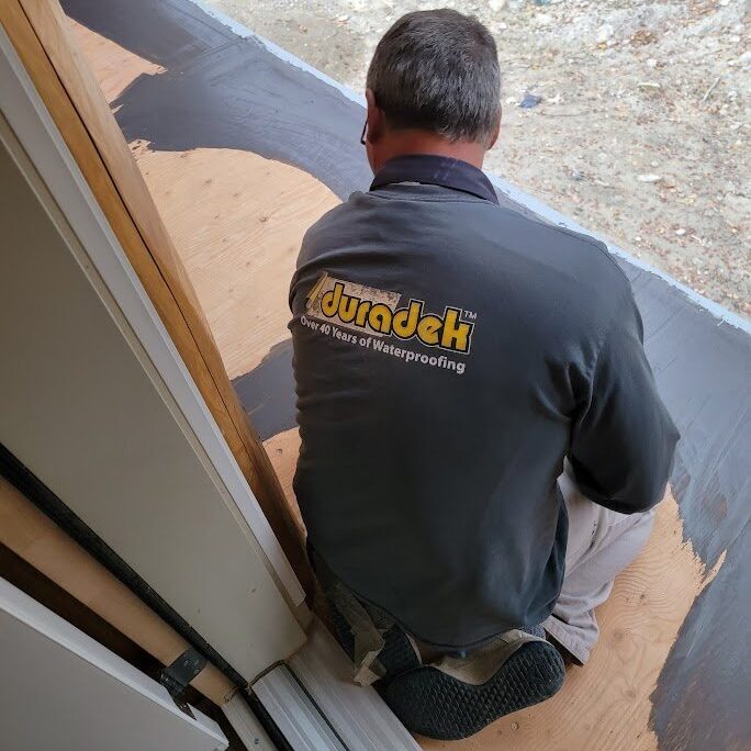 A Warwick Interiors installer applying Duradek waterproof vinyl membrane over a prepared plywood deck surface, ensuring long-lasting protection against moisture for outdoor living spaces in the Columbia Valley.